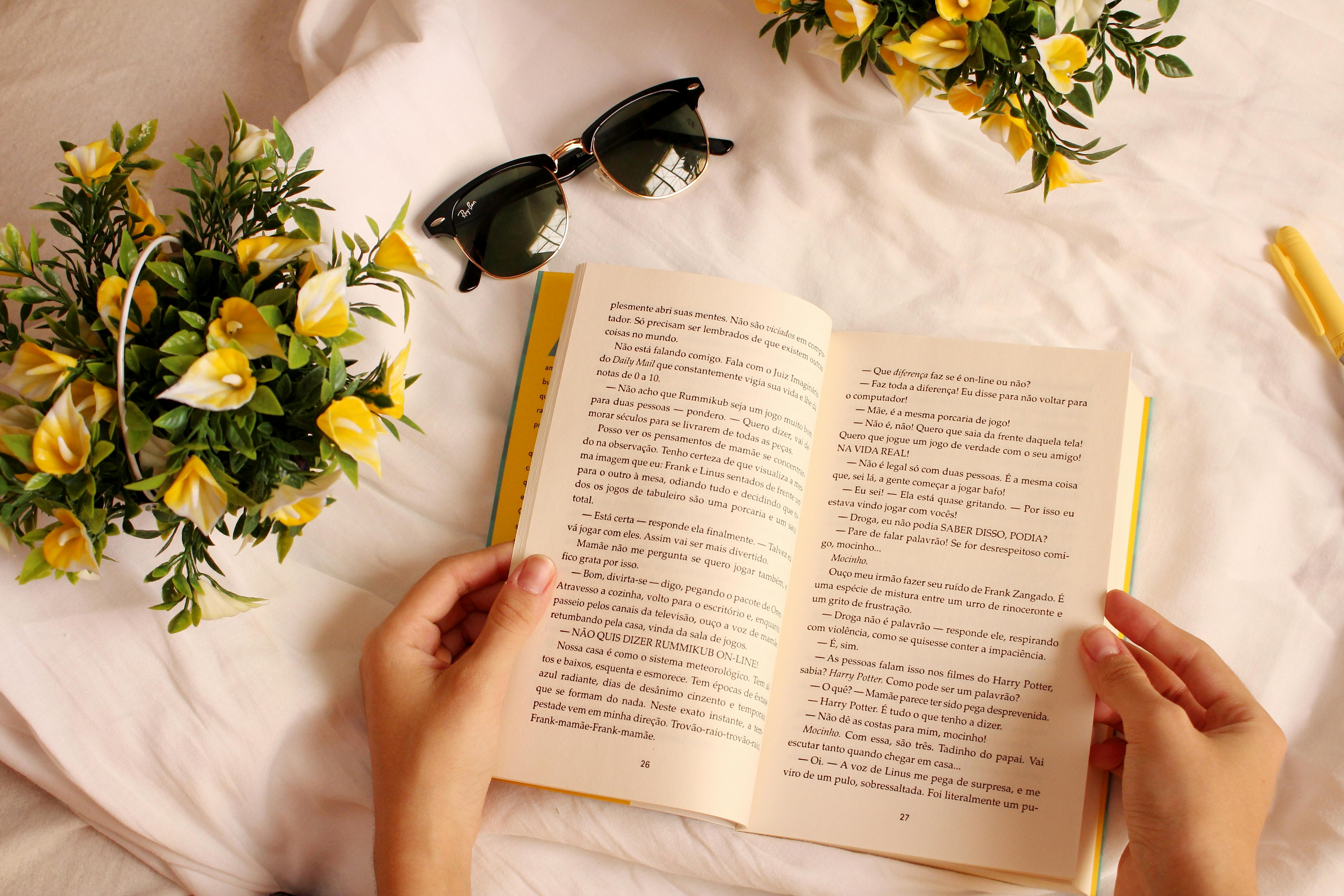 Hands holding a book surrounded by yellow flowers and sunglasses on a sunny day.